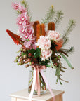 A flower arrangement in a vase sits on a wooden stool. The arrangement features Cymbidium Orchids, Roses, Banksias and Gum Foliage.