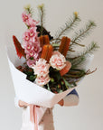 A woman holds a large flower arrangement in front of her body. The arrangement is wrapped in white paper and is tied with a pink ribbon. The arrangement features Cymbidium Orchids, Roses, Banksias and Gum Foliage.