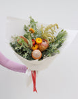 A medium size seasonal native bouquet wrapped in white paper and tied with a red ribbon. This arrangement features a protea, leucadendrons, banksias, straw flower, wattle