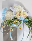 A close up of a bundle of white and green flowers. The pictured example features white/light pink roses, ammobium daisies, white gladioli, a white cymbidium orchid and a bunch of white scabiosa. Each variety of flower is wrapped in white paper individually, then wrapped again as a bundle and tied with ribbons for delivery.
