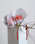 A selection of blush and pink toned flowers. Pictured are Cymbidium Orchids, Double Tulips, Blossom Branches and Erlicheer. The flowers are wrapped as a bundle in white paper with a red ribbon.