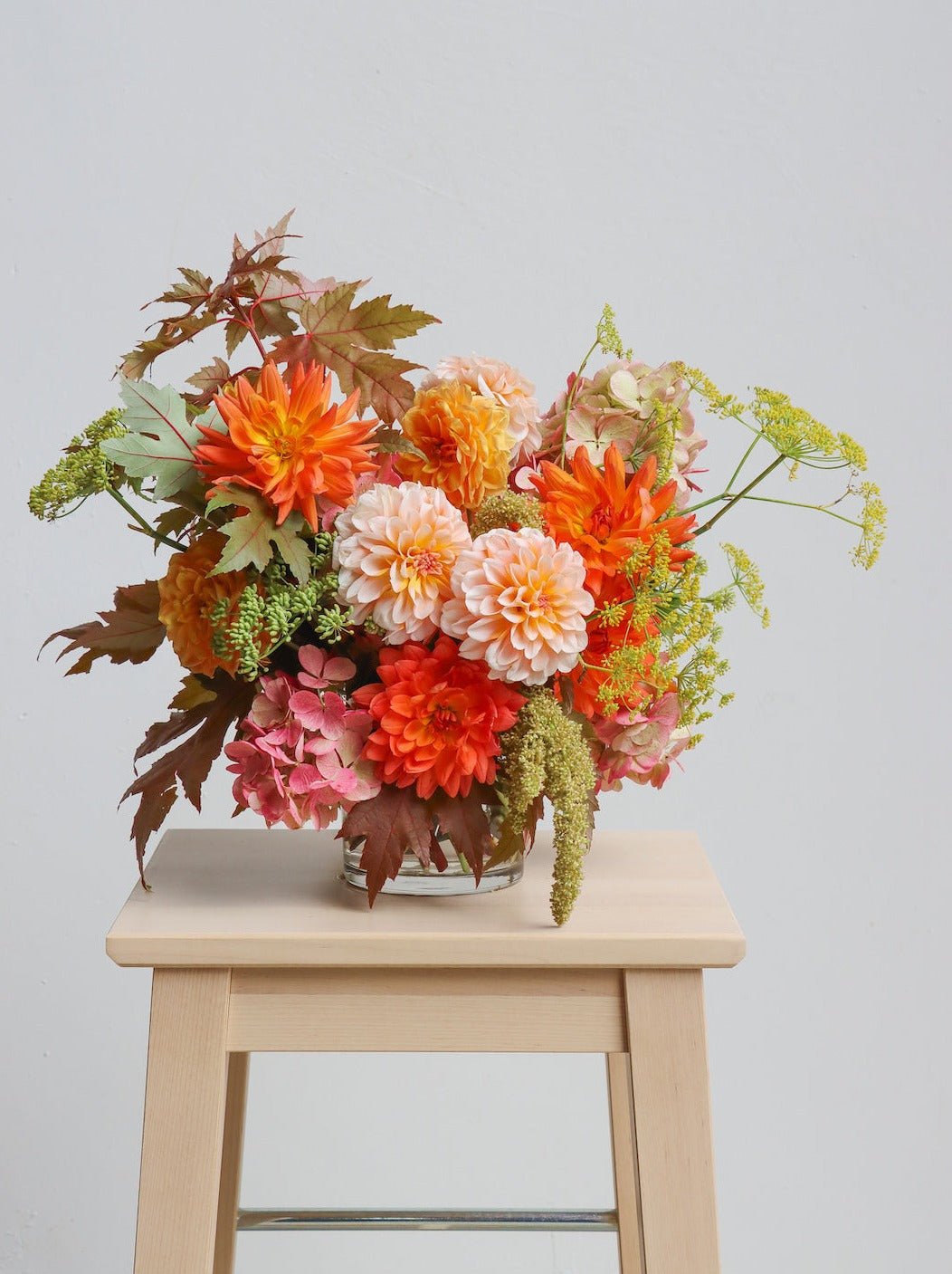 A floral arrangement in a vase featuring warm autumnal colours. The flowers/foliage pictured are Dahlias, Hydrangea, Amaranthus and Maple leaves. The arrangement is sitting on a light wooden stool in front of a white background. 