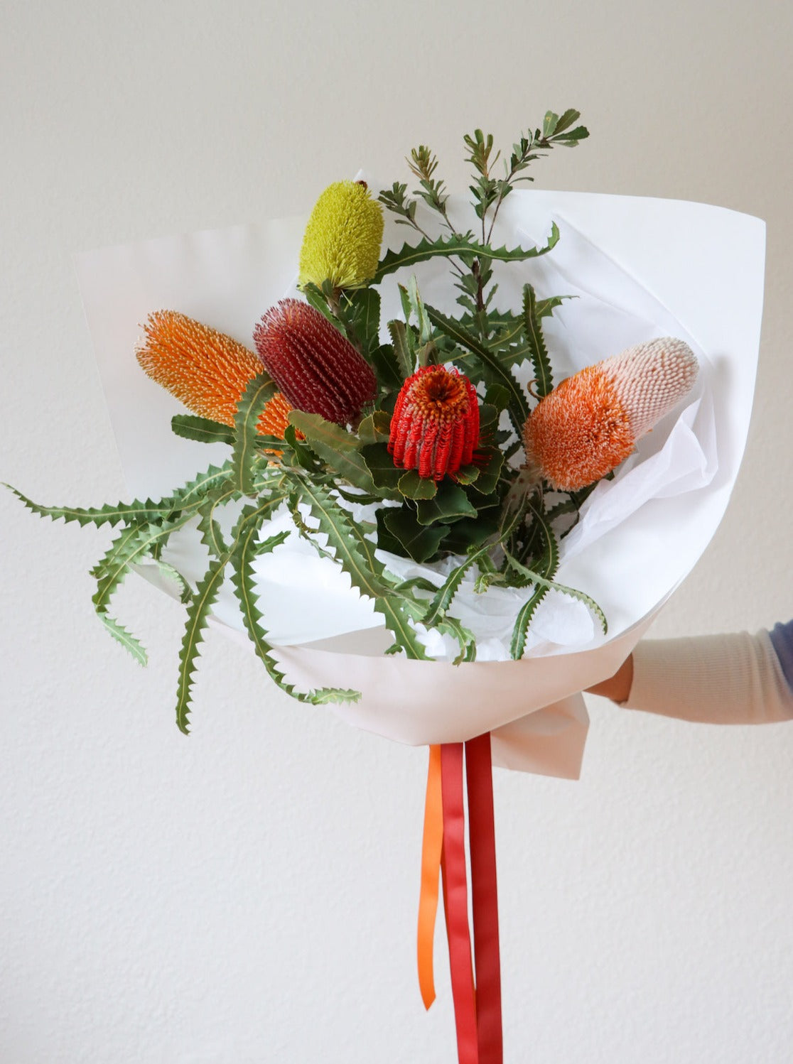 A bouquet of 5 different Banksia flowers. The Bouquet is wrapped in white paper and is tied with red and orange ribbon.