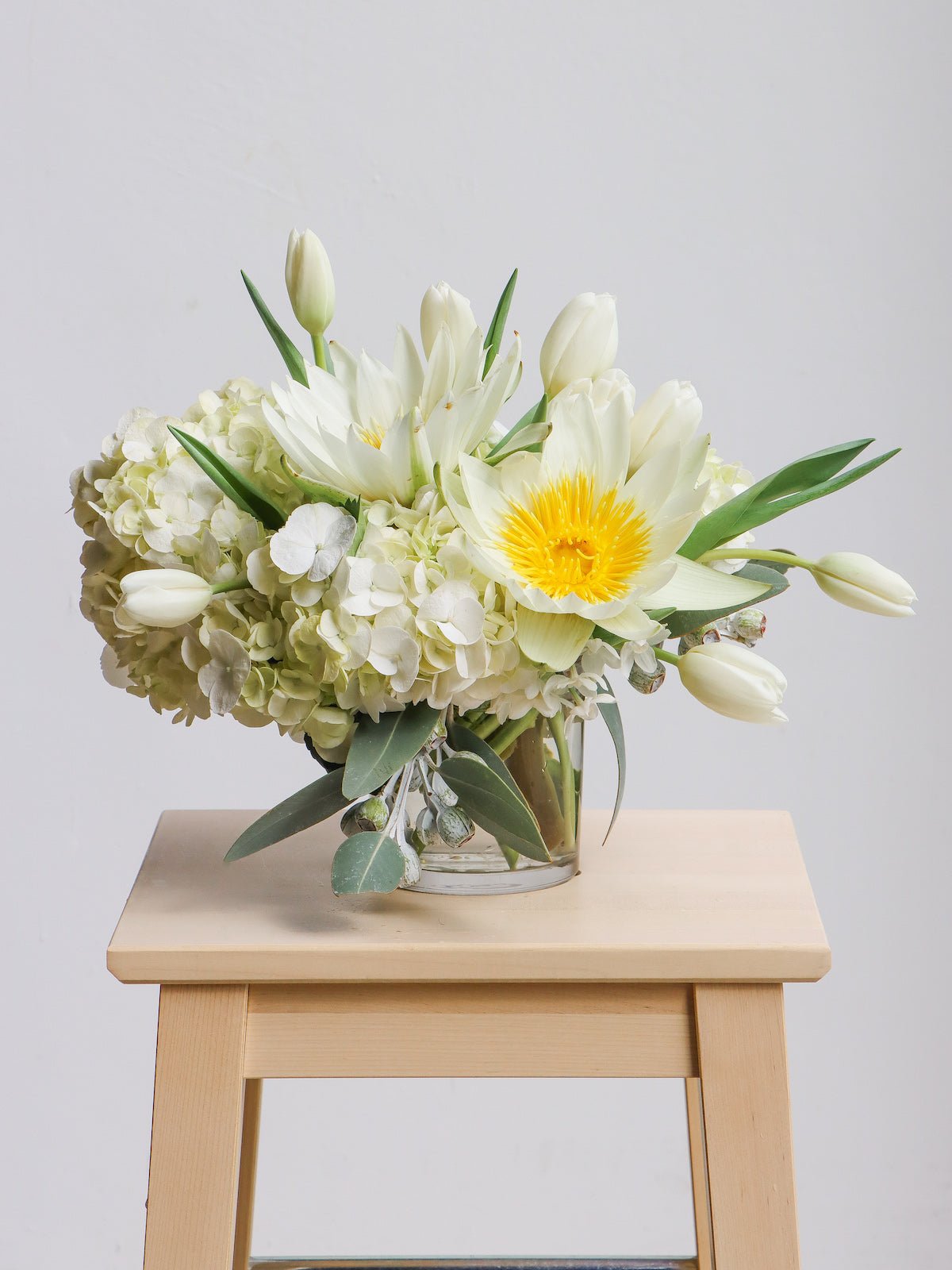 A white coloured floral arrangement in a small vase. The flowers/foliage pictured are Waterlilies, Hydrangea, Tulips and eucalyptus leaves. The arrangement is sitting on a light wooden stool in front of a white background. 
