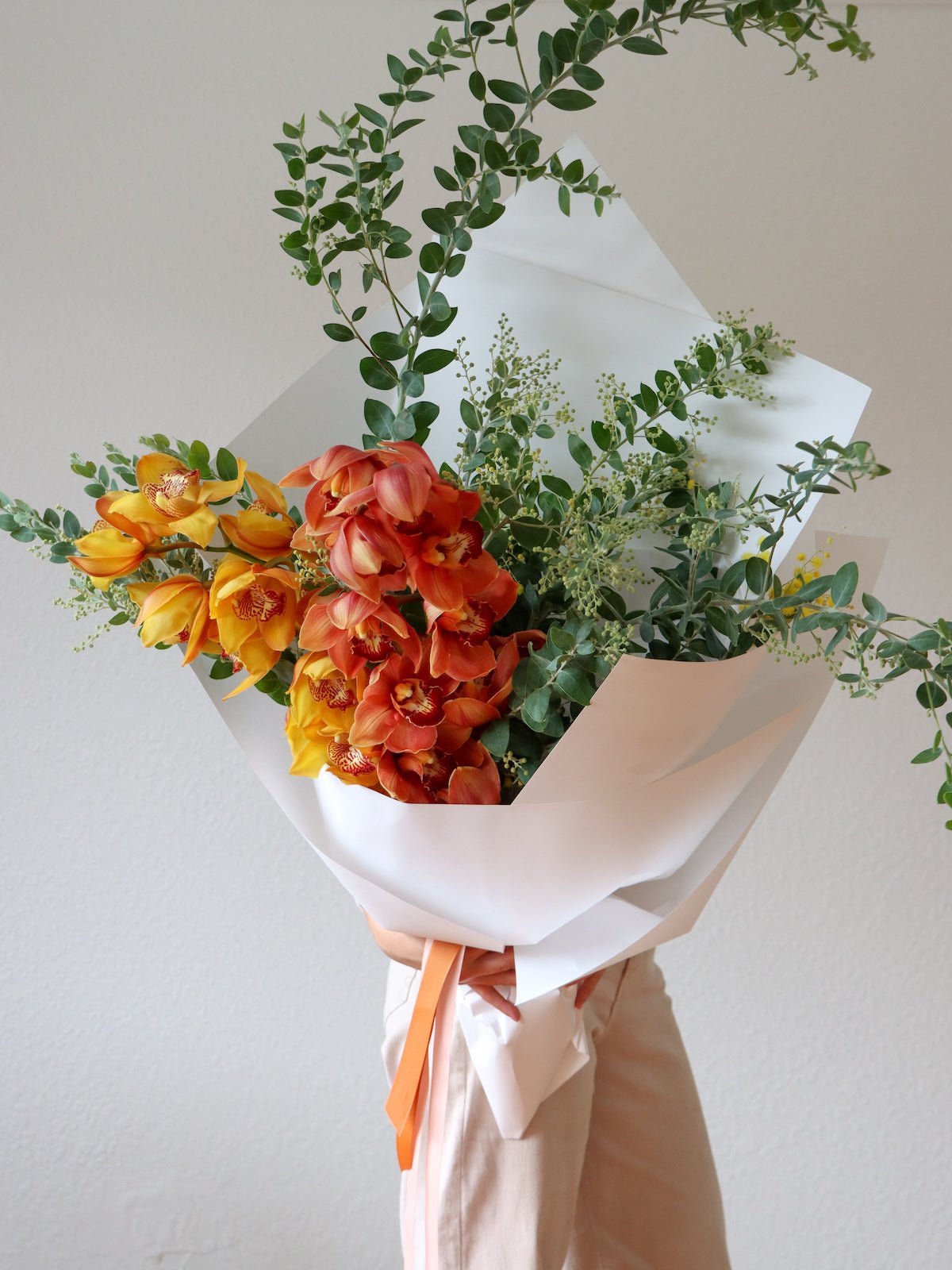 A wrapped Bouquet of 2 cymbidium orchids (one is a mustard yellow, and the other is a terracotta colour) and a silver wattle foliage with yellow flowers. The bouquet is wrapped in white paper and tied with a rust/orange coloured ribbon.  