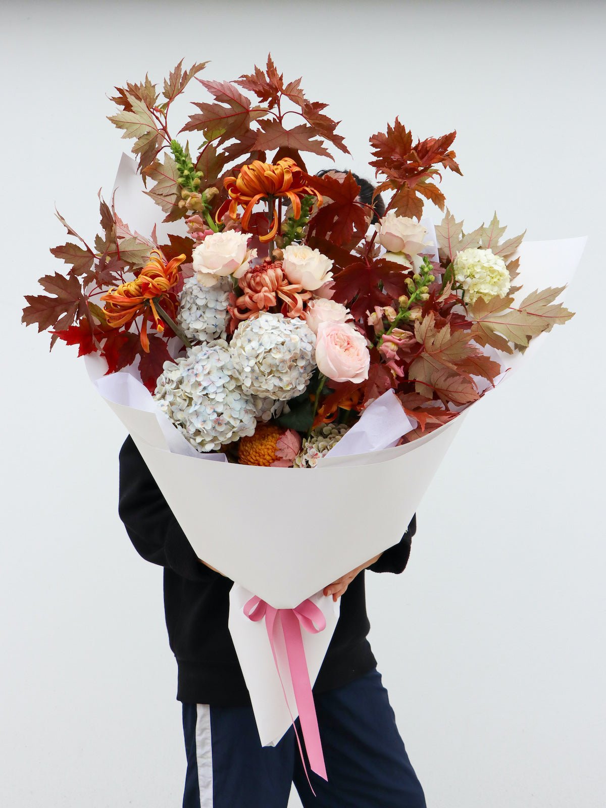 A large Autumnal toned bouquet wrapped in white paper and tied with a pink ribbon. The bouquet is held by a person in front of their body to show the large scale of the arrangement..