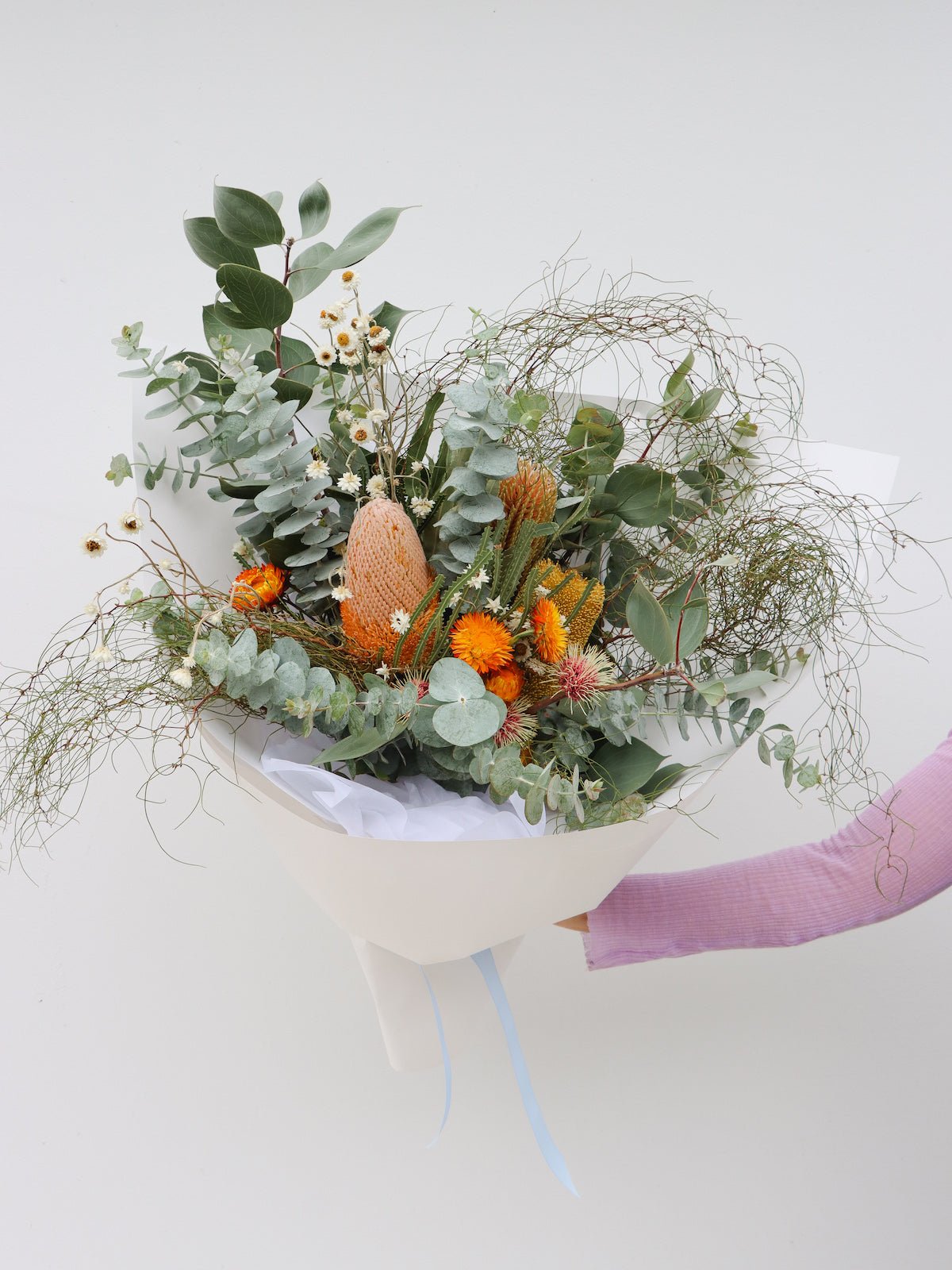 A large size seasonal native flower arrangement. The arrangement pictured features eucalyptus foliage, hakea flowers, a variety of banksias, native daisies, strawflower and zig zag wattle. The bouquet is wrapped in white paper and is tied with a baby blue ribbon. 