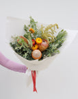 A medium size seasonal native bouquet wrapped in white paper and tied with a red ribbon. This arrangement features a protea, leucadendrons, banksias, straw flower, wattle 