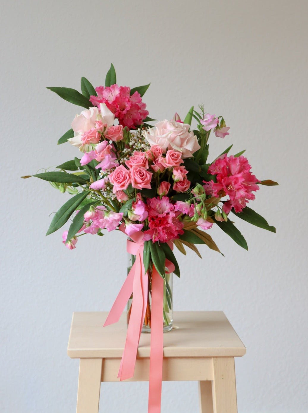 A bouquet of pink flowers sits on a wooden stool, in a cylinder glass vase. The bouquet includes pink rhododendron, pink spray roses, light pink roses and pink sweetpea. The vase has a matching pink ribbon tied around it.