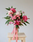 A bouquet of pink flowers sits on a wooden stool, in a cylinder glass vase. The bouquet includes pink rhododendron, pink spray roses, light pink roses and pink sweetpea. The vase has a matching pink ribbon tied around it.