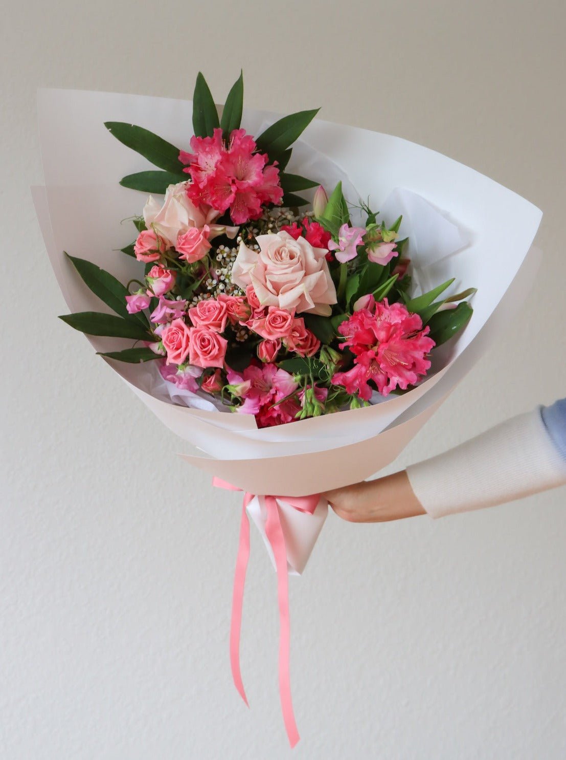 A hand is holding a bouquet of pink flowers. The bouquet includes pink rhododendron, pink spray roses, light pink roses and pink sweetpea. The bouquet is wrapped in white paper and is tied with a matching pink ribbon.