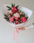 A hand is holding a bouquet of pink flowers. The bouquet includes pink rhododendron, pink spray roses, light pink roses and pink sweetpea. The bouquet is wrapped in white paper and is tied with a matching pink ribbon.