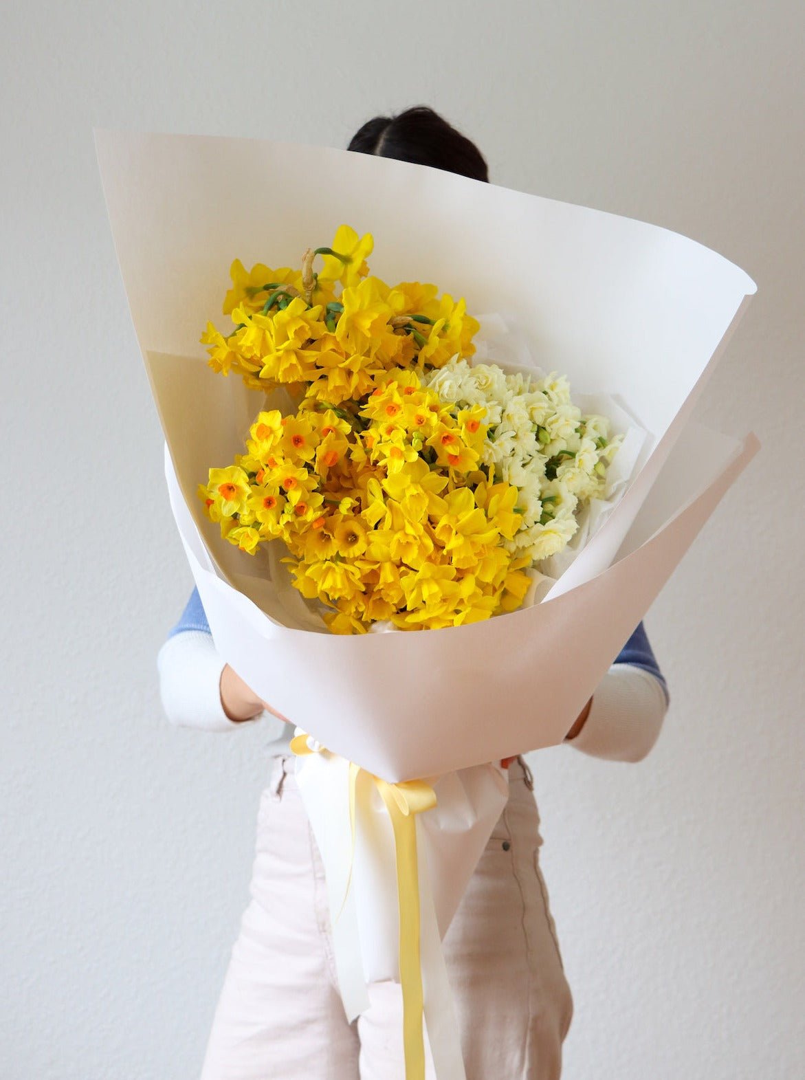 holding a fragrant bouquet of seasonal Narcissus flowers, including Daffodils and Erlicheer, wrapped in white paper.