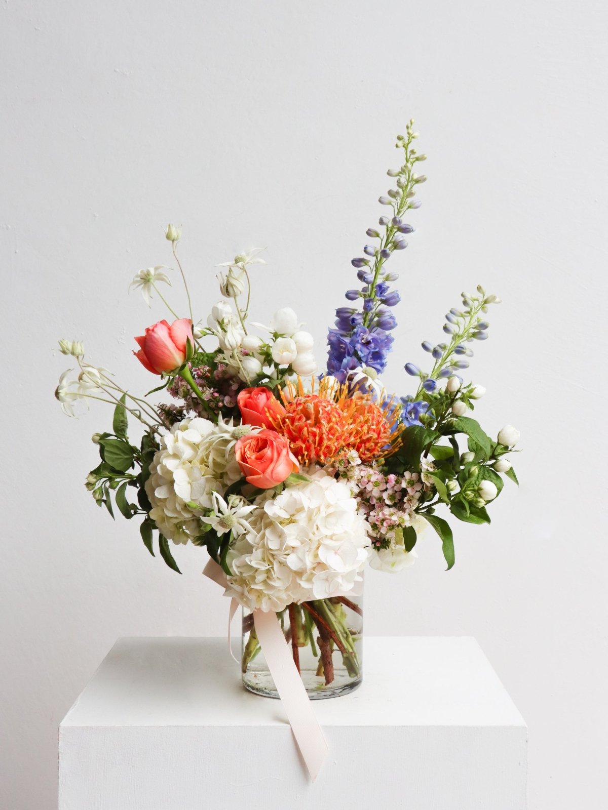 A garden style arrangement in a glass vase. The arrangement features delphinium, hydrangea, pincushions, roses, mock orange and flannel flowers. The arrangement is sitting on a white plinth in front of a white wall.