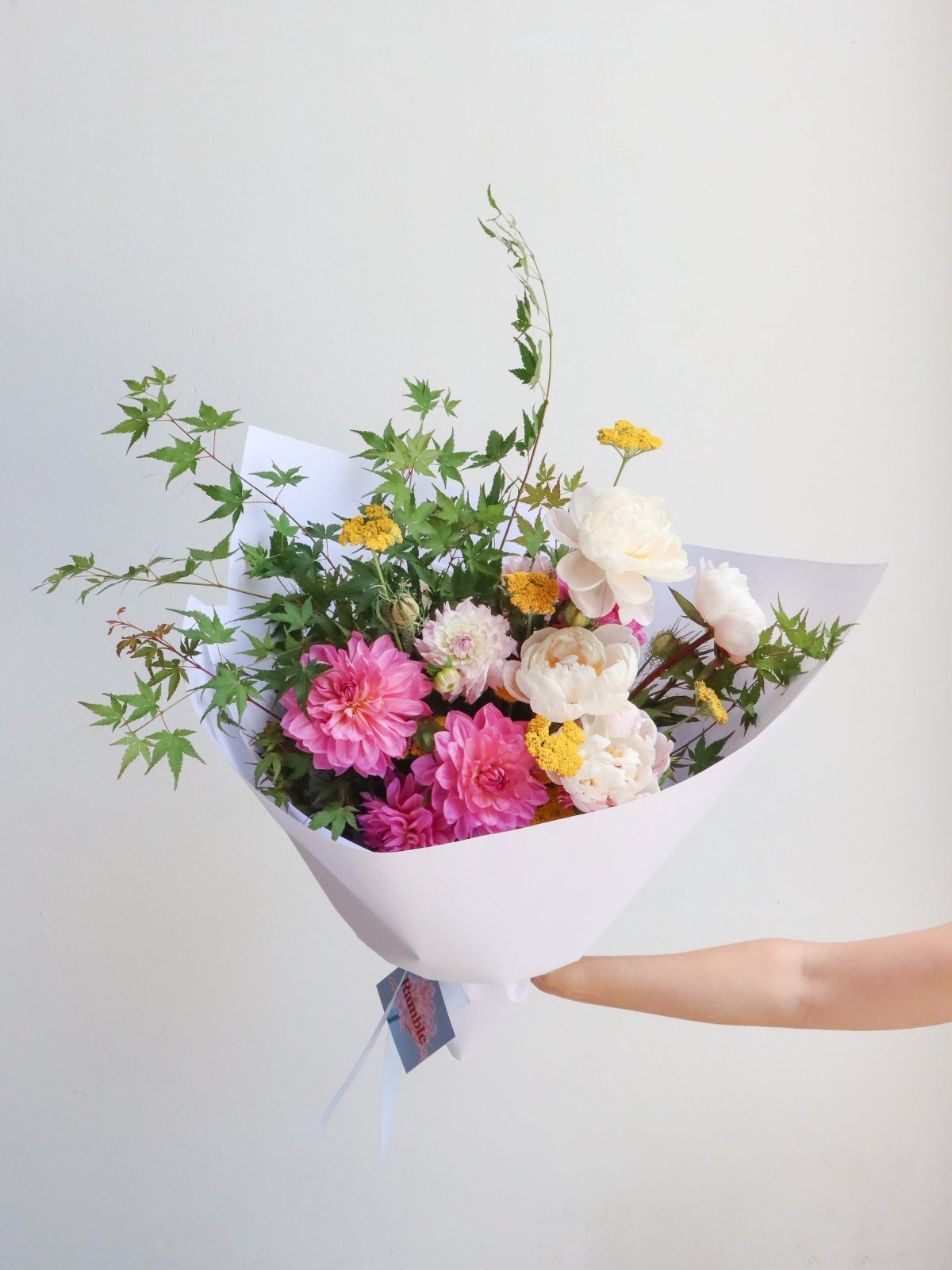 A bouquet of seasonal flowers, wrapped in white paper. The example pictured features bright pink dahlias, light pink peonies, maple foliage, and yellow achillea. 